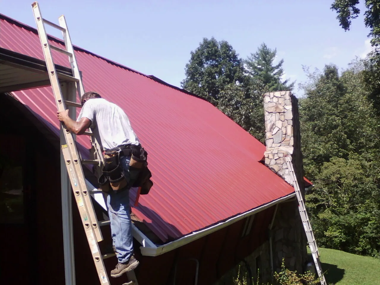 Roofer on a ladder beside a red metal roof and stone chimney — Soffit & Fascia Repair by Roofing Brothers in New Century
